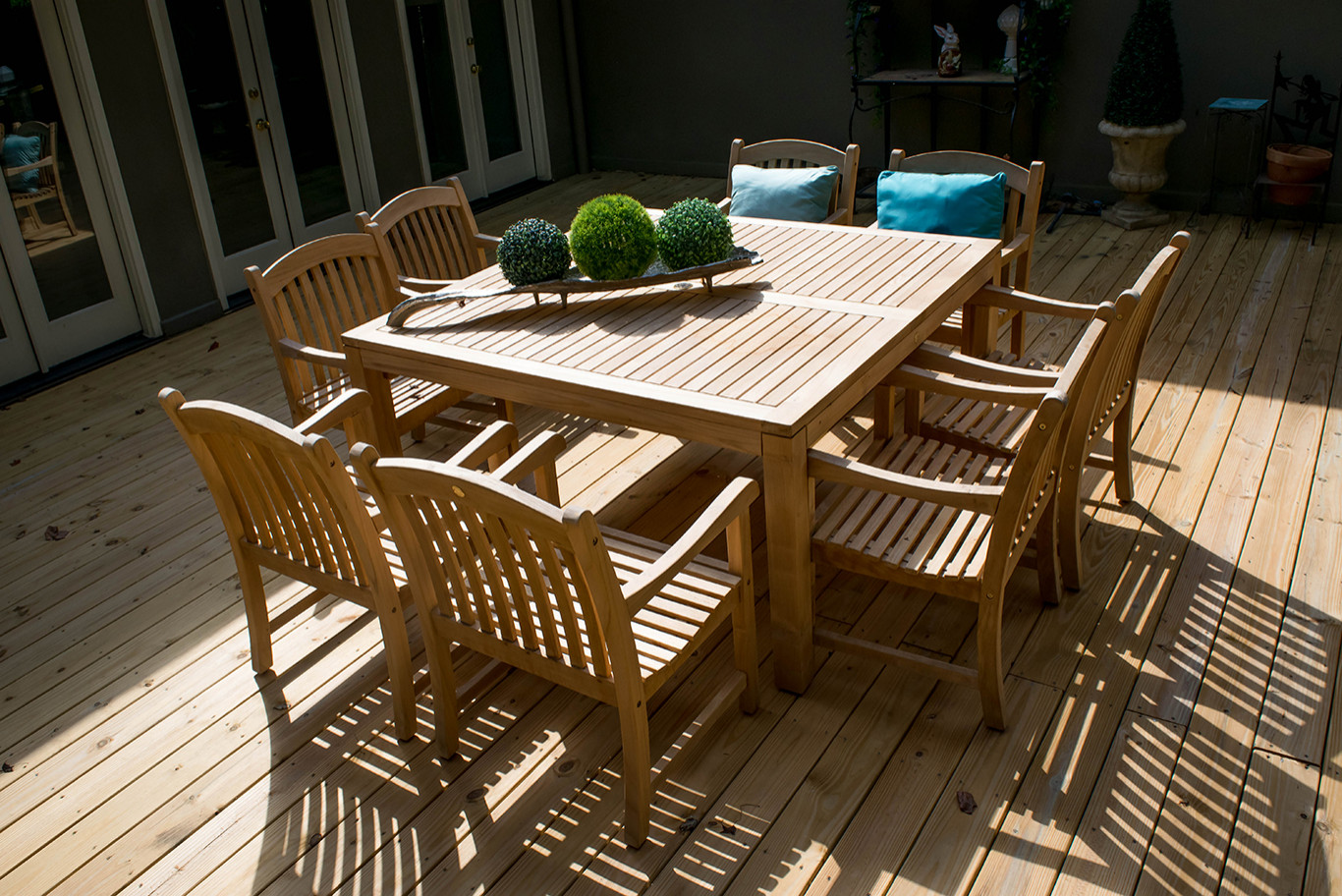 A wooden outdoor dining table with eight matching chairs on a wooden deck, decorated with a rectangular tray holding three spherical plants.