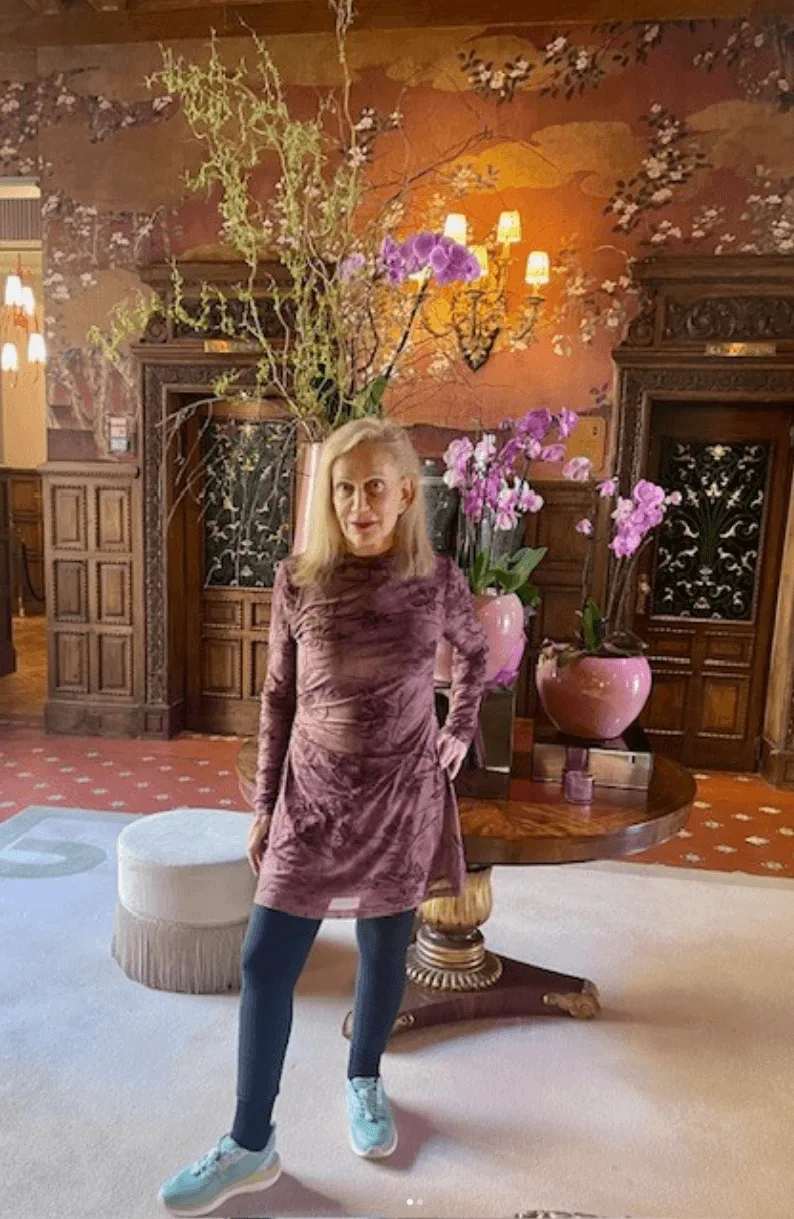 About Linda Rickels: A woman in a purple dress and blue sneakers stands in front of a round table with pink vases and orchids, in an ornately decorated room with wood paneling and floral wallpaper.