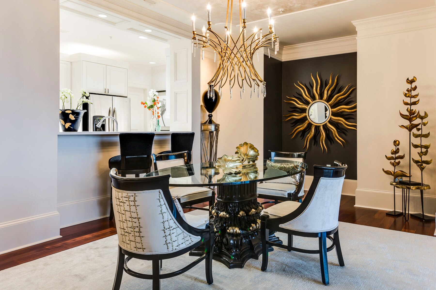 A dining room with a round glass table, four chairs, a modern gold chandelier, sunburst wall clock, and decorative metallic tree sculptures. The kitchen is visible in the background.