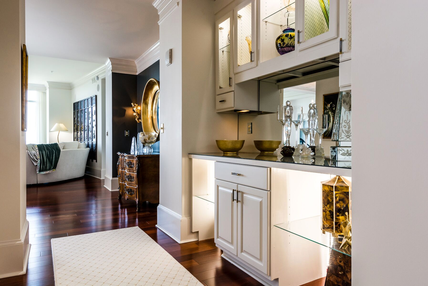 A modern hallway with a built-in glass and white cabinet, decorative items on shelves, hardwood floors, and a view into a living room with a sofa and artwork, showcasing the kind of spaces included in our design services.