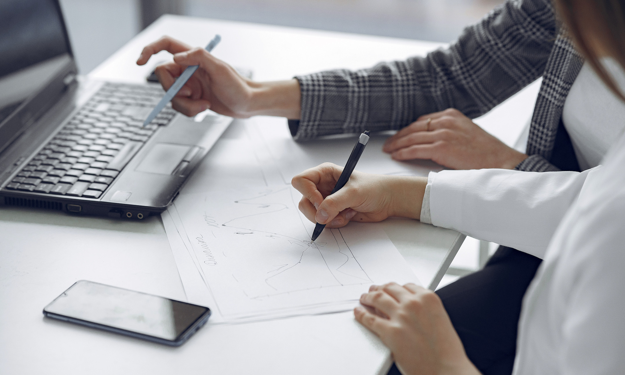 Two people are reviewing printed charts and graphs on paper at a desk, using pens and referencing a laptop and a smartphone, possibly analyzing client testimonials.