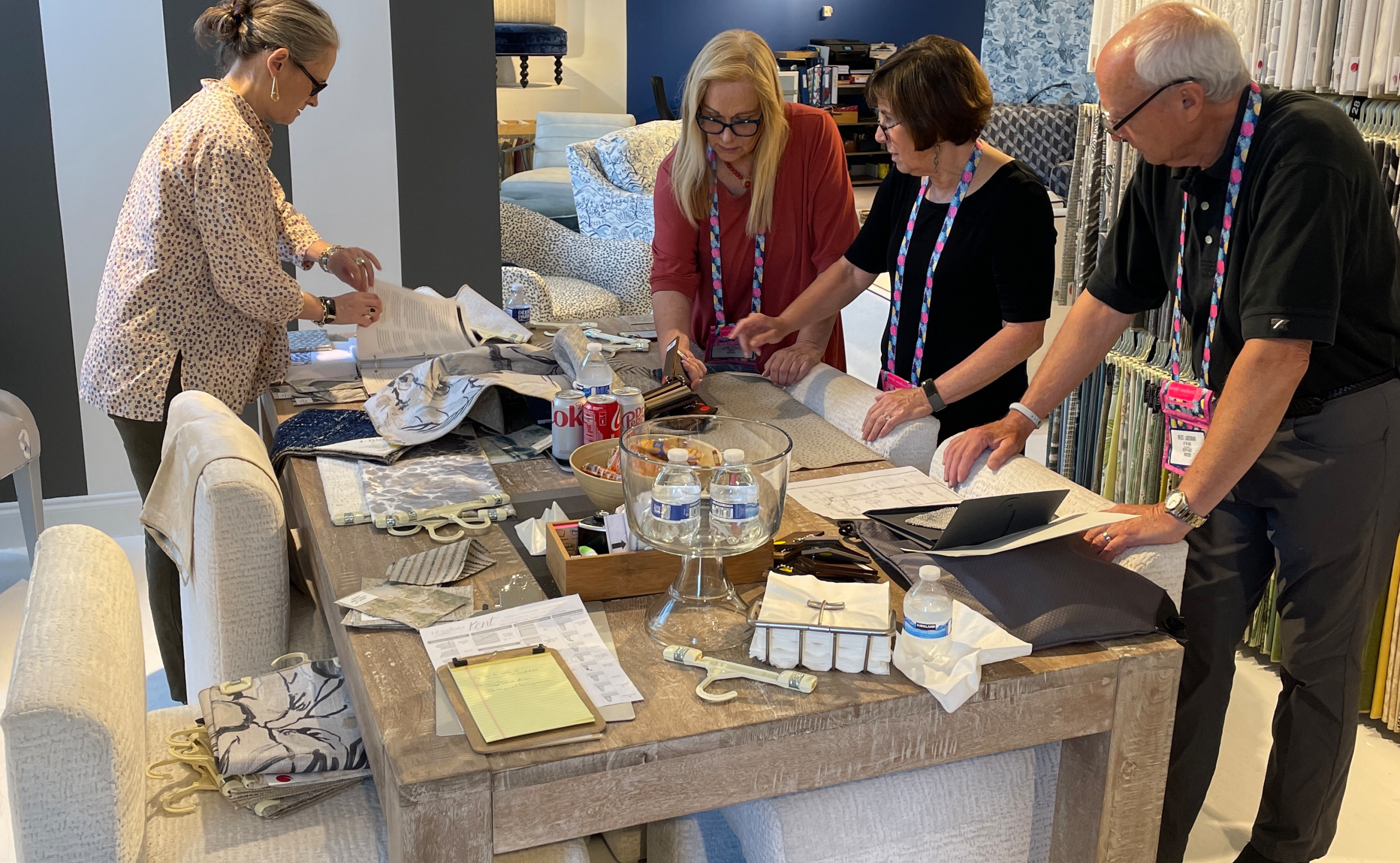 Four people stand around a large table covered with fabric samples, papers, and drinks, working collaboratively in a showroom or design studio as part of the interior design process.