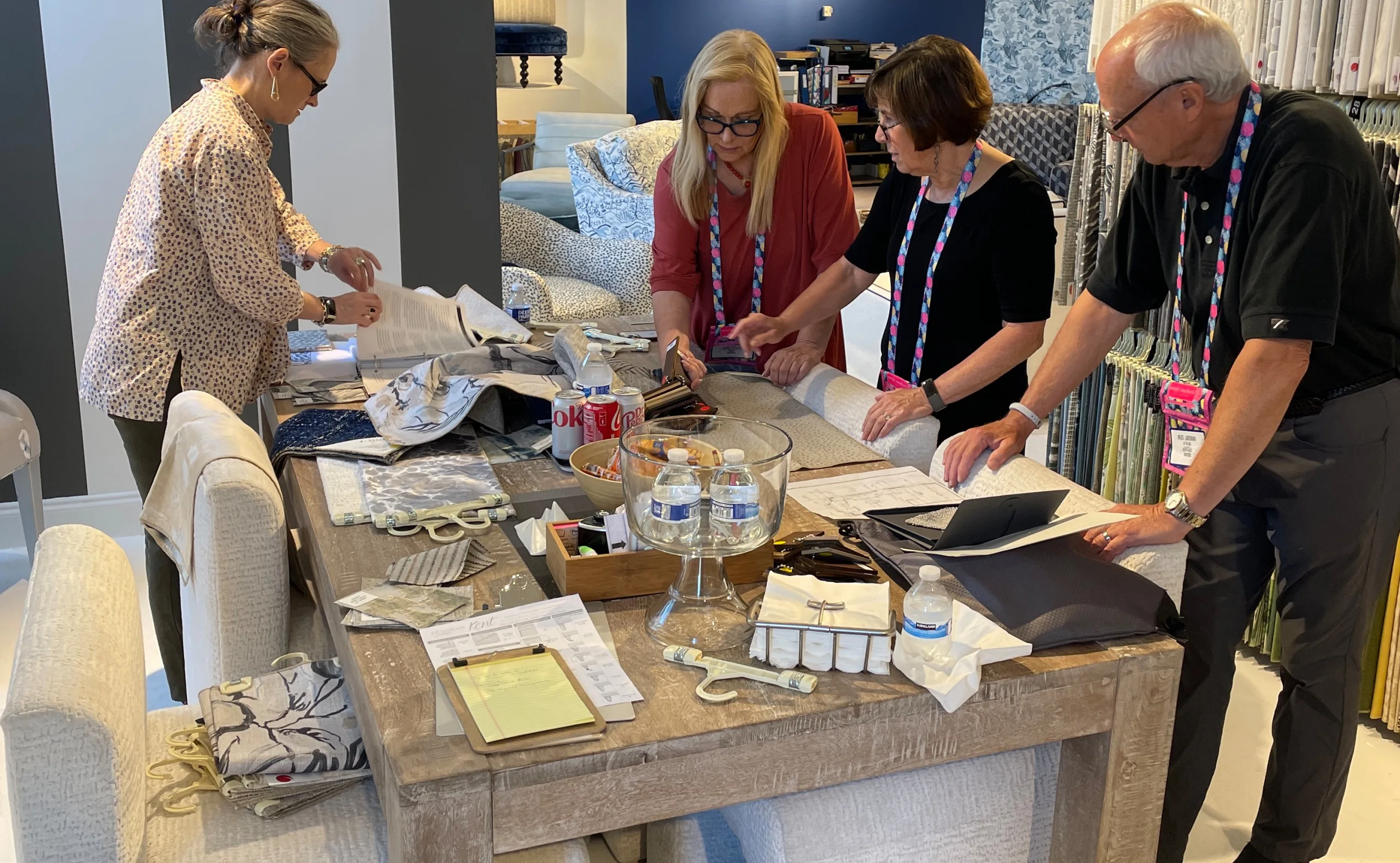 Four people stand around a large table covered with fabric samples, papers, and drinks, working collaboratively in a showroom or design studio as part of the interior design process.