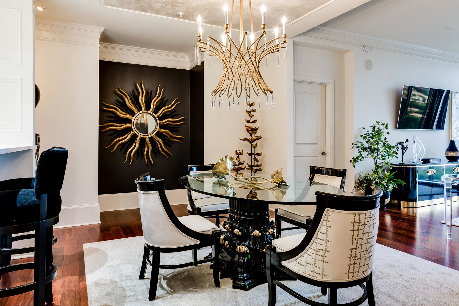 Modern dining room with a glass round table, four upholstered chairs, gold chandelier, sunburst wall art, and contemporary decor on hardwood floor.
