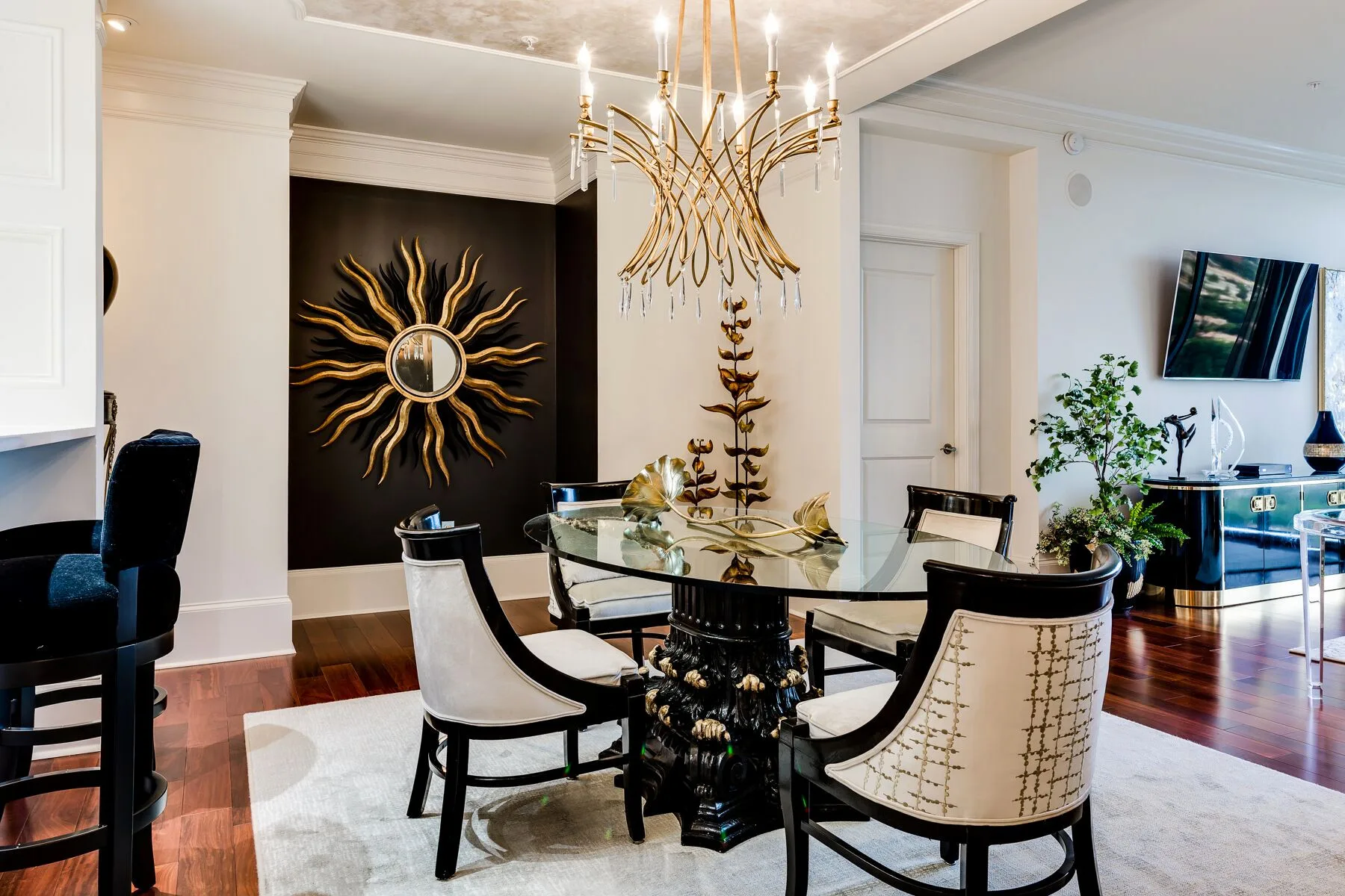 Modern dining room with a glass round table, four upholstered chairs, gold chandelier, sunburst wall art, and contemporary decor on hardwood floor.
